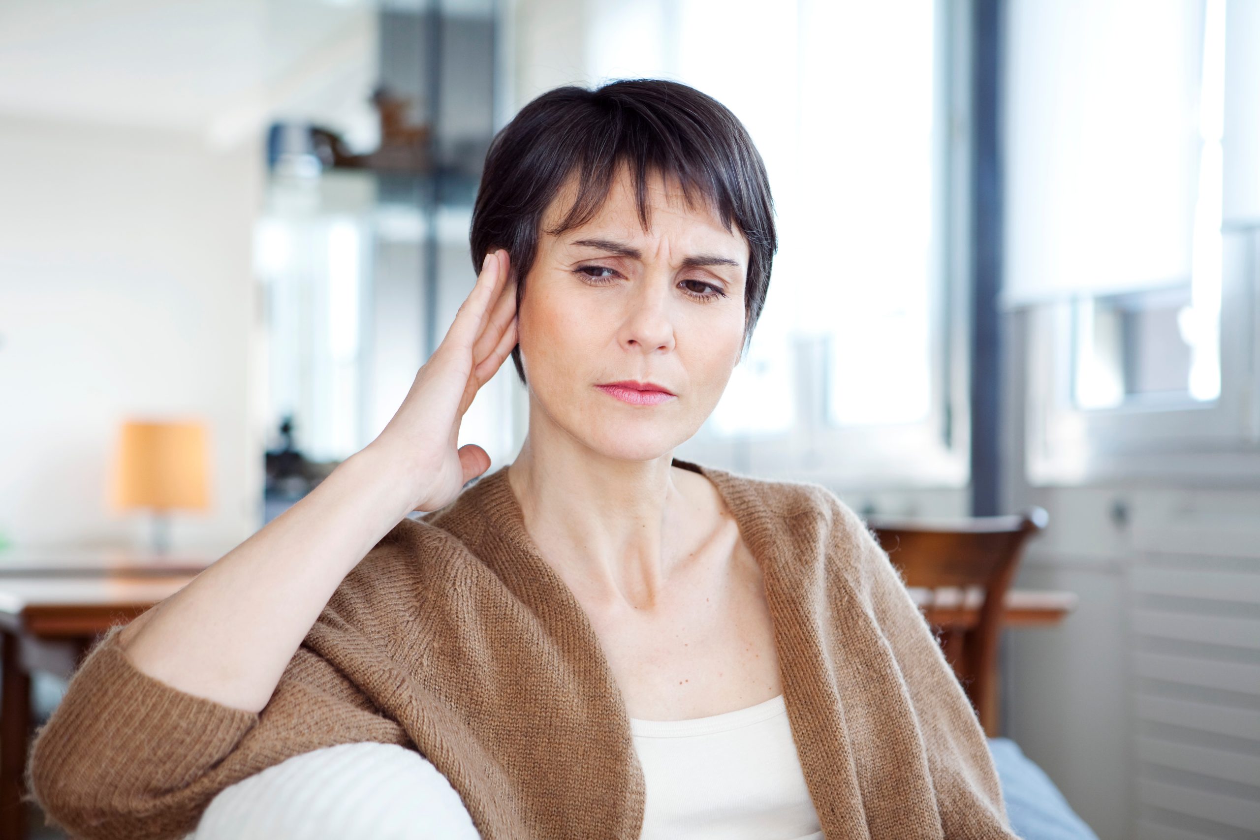 Eine Frau mit kurzen dunklen Haaren hält sich nachdenklich und mit einer Hand ans Ohr, was auf Ohrgeräusche oder Tinnitus hindeutet. CMD kann als Ursache solcher Beschwerden eine Rolle spielen.