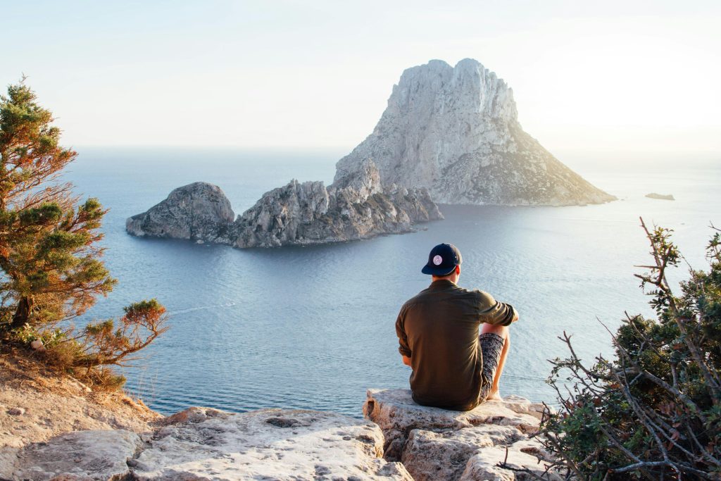 Ein Mann sitzt auf einem Felsen und blickt auf eine malerische Küstenlandschaft. Diese entspannte Szene symbolisiert die Wichtigkeit von Pausen und Entspannung für Menschen mit CMD.