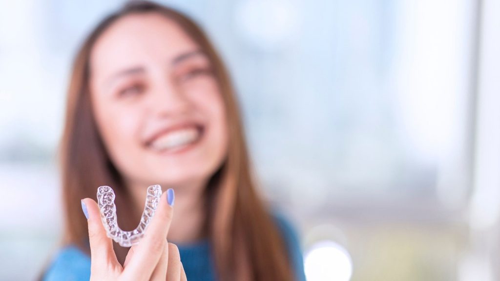 Junge Frau hält einen transparenten Aligner in der Hand und lächelt. Diese moderne kieferorthopädische Lösung wird häufig bei CMD eingesetzt, um Fehlstellungen der Zähne sanft zu korrigieren. Aligner bieten eine diskrete und flexible Möglichkeit, die Kieferposition zu verbessern und CMD-Symptome zu lindern.