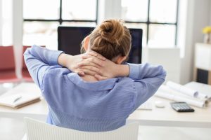 Frau am Schreibtisch, die sich mit beiden Händen den verspannten Nacken hält, ein typisches Symptom bei CMD.