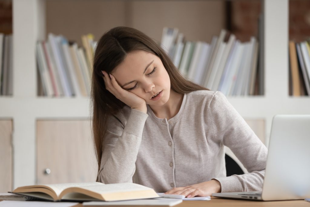 Eine Frau mit langen braunen Haaren sitzt erschöpft am Schreibtisch mit einem offenen Buch und Laptop, möglicherweise wegen Schlafmangel durch CMD. Ihre müde Haltung zeigt die Auswirkungen von Schlafstörungen und CMD auf die Konzentration und Energie.