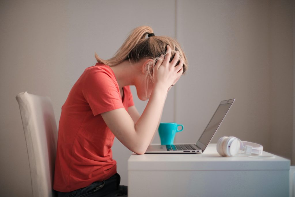 Eine gestresste Frau sitzt vor einem Laptop und hält ihren Kopf in den Händen. Dieses Bild symbolisiert die negativen Auswirkungen von Stress, der als Auslöser oder Verstärker von CMD-Symptomen wie Kieferschmerzen wirken kann.