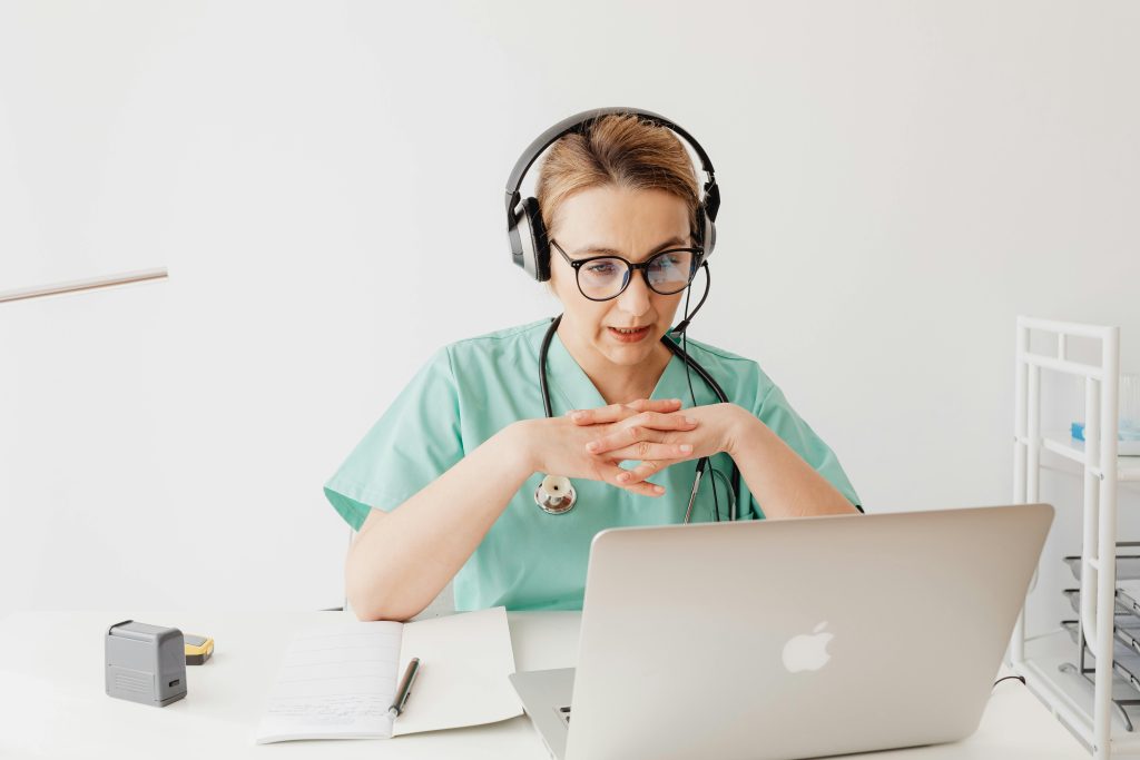 Eine Ärztin in grüner Kleidung und mit einem Stethoskop sitzt an einem Schreibtisch und führt eine Online-Beratung durch. Sie trägt ein Headset und spricht konzentriert in die Kamera eines Laptops.