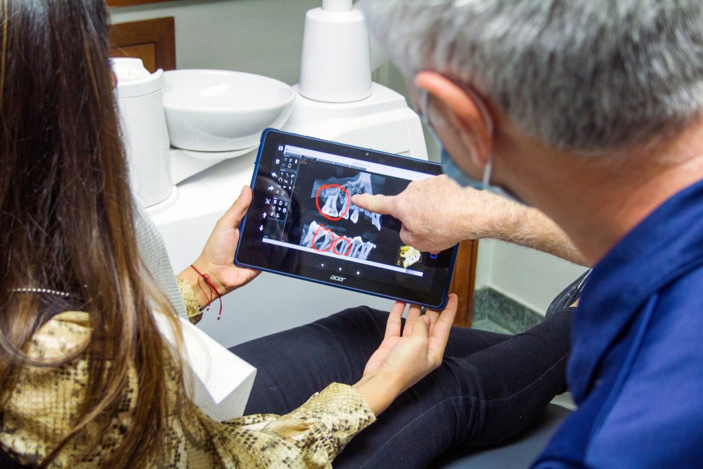 Eine Patientin und ein Zahnarzt betrachten Röntgenbilder auf einem Tablet. Der Zahnarzt zeigt auf markierte Zahnfehlstellungen, die mögliche Ursachen für CMD sein könnten.