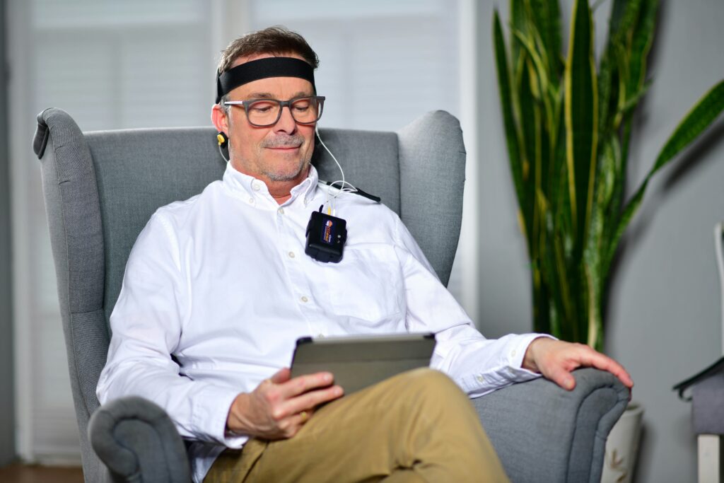 Ein Mann entspannt in einem Sessel während einer Biofeedback-Therapie. Er trägt ein Kopfband mit Sensoren und ein tragbares Biofeedback-Gerät, das visuelle Rückmeldungen liefert. Diese Methode hilft, Muskelverspannungen bei CMD gezielt zu kontrollieren.