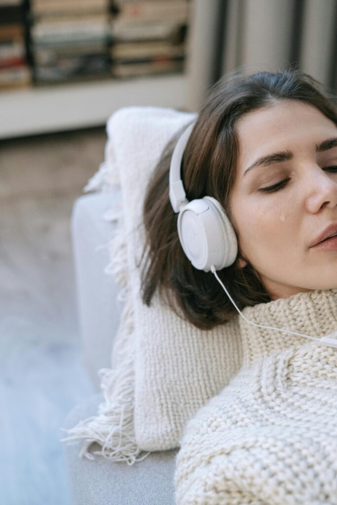 Entspannte Frau auf einem Sofa, eingehüllt in eine Decke, während sie beruhigende Klänge über Kopfhörer hört. Musiktherapie kann eine wohltuende Ergänzung zur CMD-Behandlung sein, um die Kaumuskulatur zu entspannen und Schmerzen zu lindern.