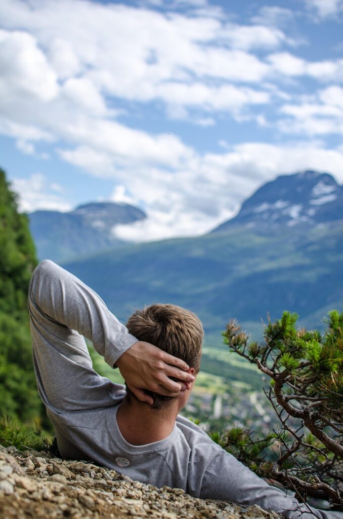 Ein Mann liegt entspannt auf einem Hügel mit Blick auf eine malerische Berglandschaft. Die Ruhe der Szene vermittelt den positiven Effekt von Auszeiten auf CMD-Beschwerden.