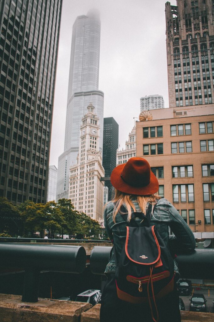 Eine Frau mit einem roten Hut betrachtet die Skyline einer Stadt, während sie einen Rucksack trägt. Die Haltung zeigt Entspannung, ideal für Themen wie CMD und Reisen oder stressfreies Erkunden.