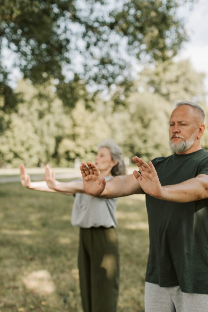 Ein älteres Paar steht in einer natürlichen Parklandschaft und führt typische Tai Chi Bewegungen aus. Die ausgestreckten Hände und der achtsame Ausdruck verdeutlichen die kontrollierte, harmonische Bewegung, die das Gleichgewicht fördert. Diese Praxis hilft CMD-Betroffenen, Fehlhaltungen zu korrigieren und die Belastung auf das Kiefergelenk zu minimieren. Die ruhige Umgebung im Freien trägt zur mentalen und körperlichen Entspannung bei, was die Heilung nachhaltig unterstützen kann.