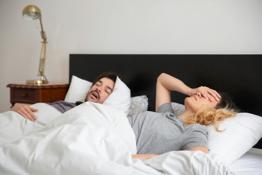 Mann schnarcht im Bett, während seine Partnerin genervt die Hand auf die Stirn legt – Symbolbild für nächtliche Atemstörungen und Schnarchprobleme durch CMD.